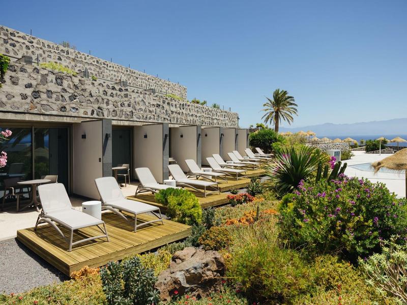 Modern hotel rooms with lounge chairs, surrounded by tropical plants and sea view.