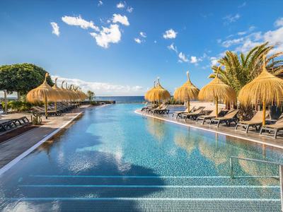 Vue d'une longue piscine avec des transats et des parasols en paille, la mer en arrière-plan sous un ciel ensoleillé.