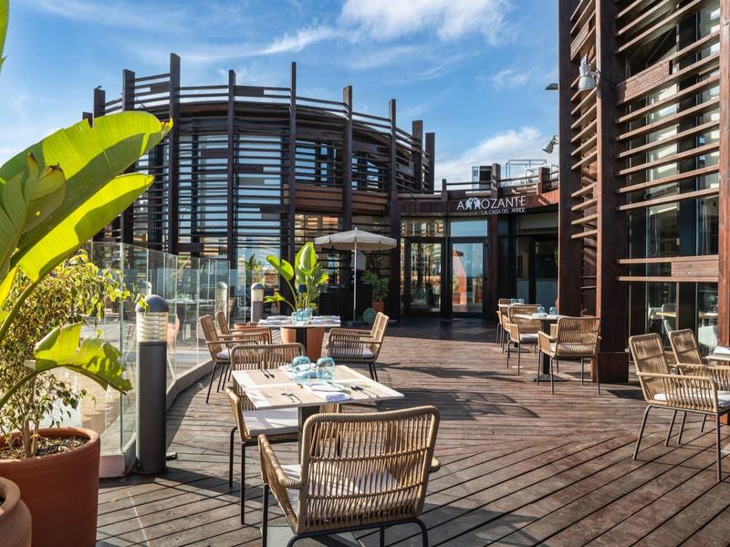 Área exterior moderna de hotel con suelo de madera, mesas, sillas y plantas bajo cielo azul.