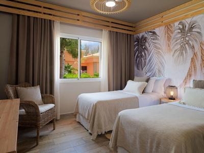 Habitación de hotel con dos camas individuales, silla, ventana con vista al exterior y decoración mural tropical.