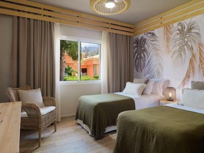 Habitación de hotel con dos camas, decoración de pared temática de palmeras y vista por la ventana.