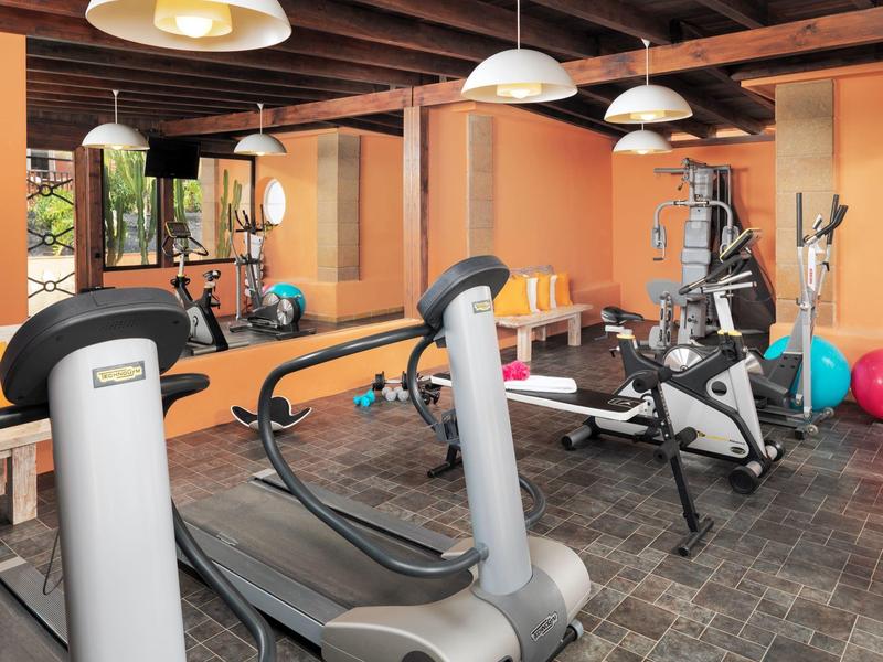 Ein helles Trainingszimmer mit Laufband, Fahrradtrainer, Spiegeln und orangener Wandgestaltung.