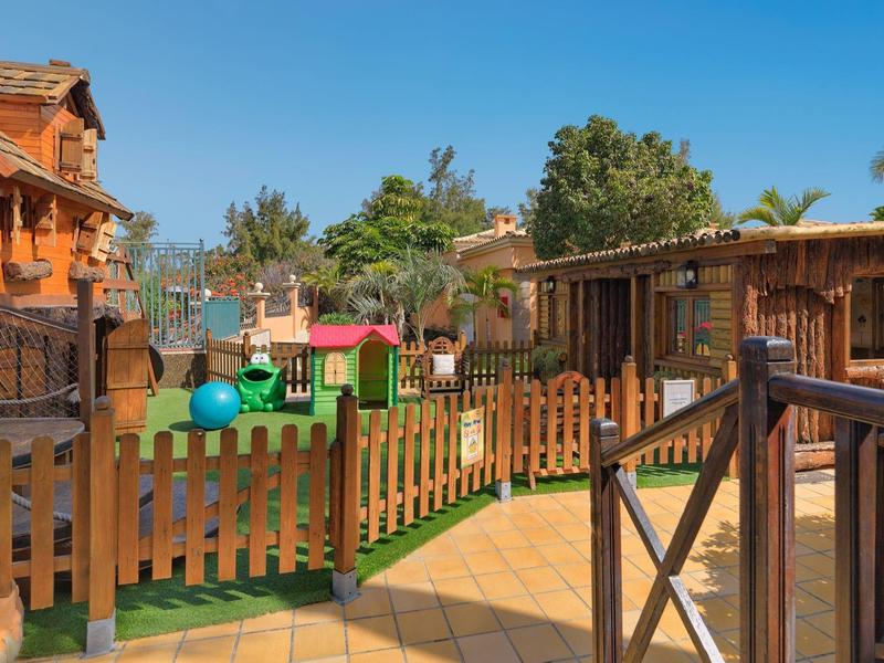 Kinderspielplatz mit Holzhäusern, grünem Gras, bunten Spielgeräten und Holzzäunen bei sonnigem Wetter.