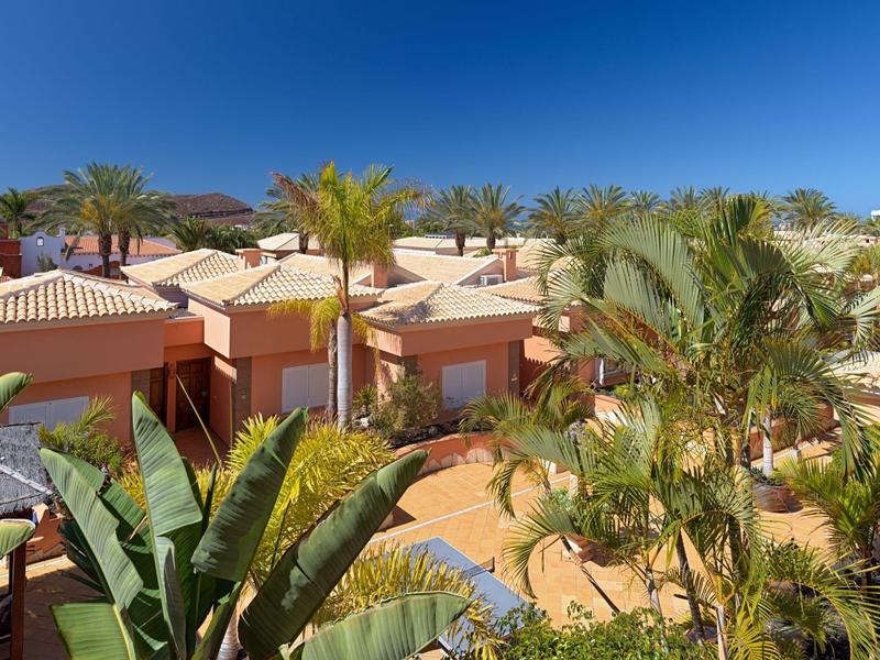 Wohnhäuser mit roten Dächern, umgeben von Palmen und Pflanzen unter klarem blauem Himmel.