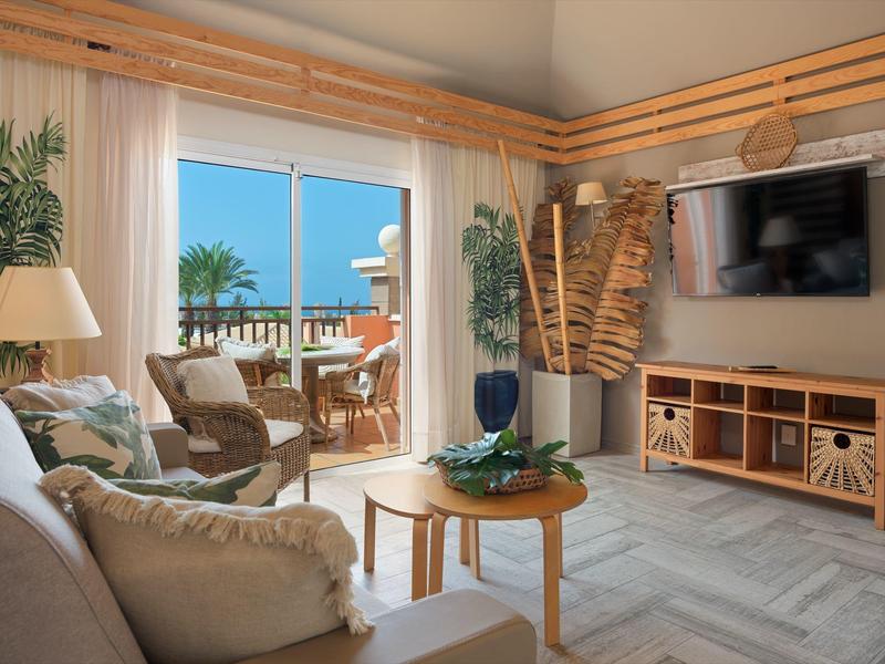 Gemütliches Wohnzimmer mit Holzmöbeln, großen Fenstern und Blick auf Terrasse und Meer.