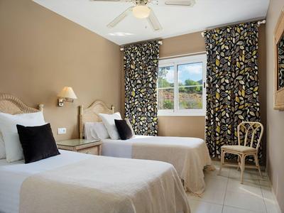 Camera d'hotel con due letti singoli, pareti beige, tende a motivo e una sedia vicino alla finestra.