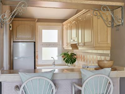 Cucina compatta con mobili in legno chiaro, lavello, frigorifero e due sedie al bancone.
