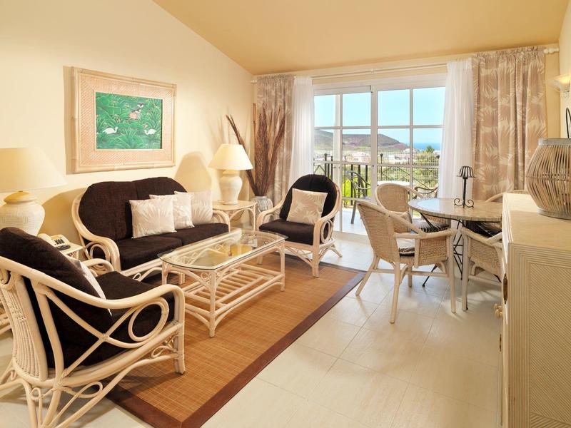 Helles Wohnzimmer mit Korbmöbeln, Glas-Couchtisch, Teppich und großem Fenster mit Blick ins Freie.