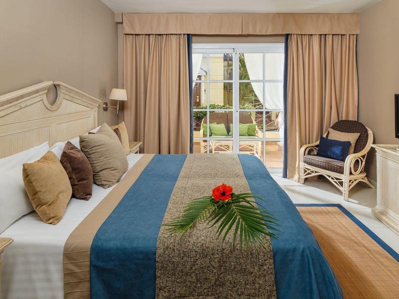 Hell eingerichtetes Schlafzimmer mit großem Bett, blauen Läufern und Terrasse im Hintergrund.