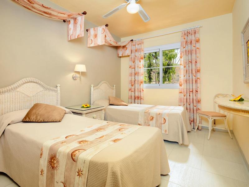 Hell eingerichtetes Schlafzimmer mit zwei Einzelbetten, Fenstervorhängen und kleinem Schreibtisch.