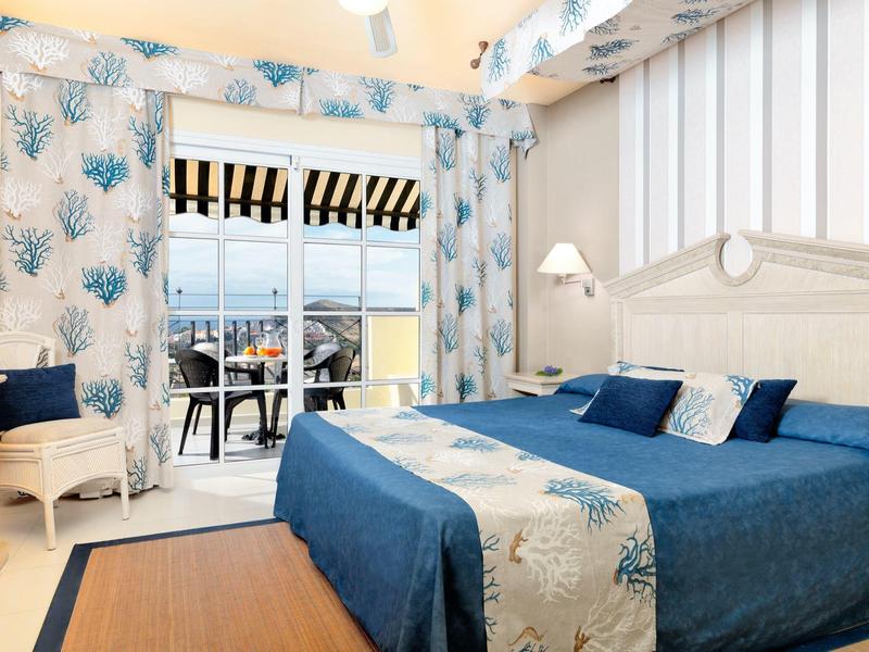 Helles Schlafzimmer mit Blautönen, floralem Muster, Doppelbett und Balkon mit Meerblick.