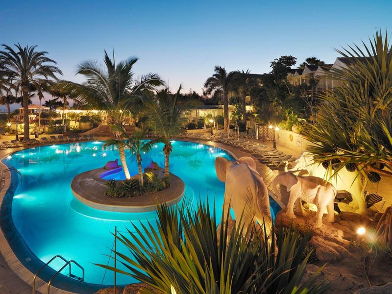 Beleuchteter Pool mit Insel und Palmen, umgeben von Pflanzen und Deko mit Giraffenfiguren.