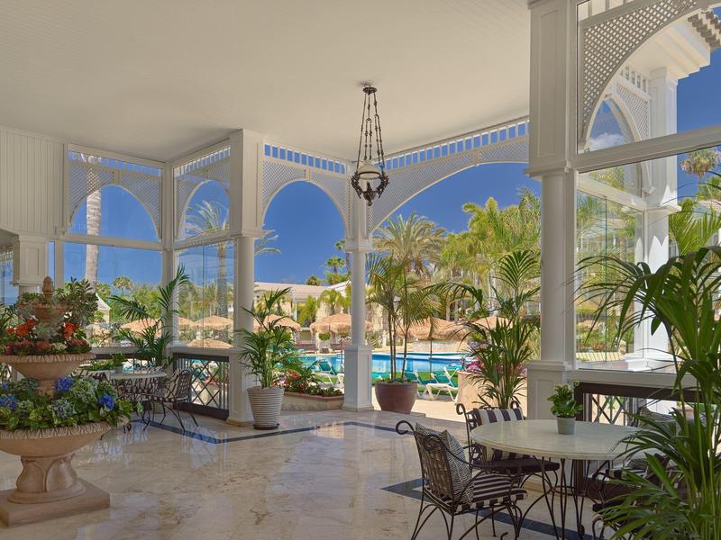 Offener Wintergarten mit weißen Bögen, Sitzgruppen und Blick auf Pool und Palmen bei blauem Himmel.