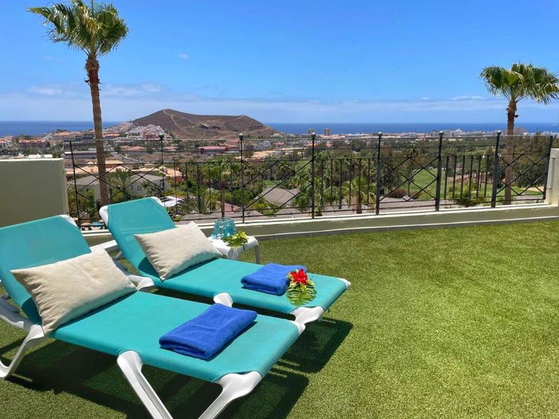 Zwei türkisfarbene Liegen mit Kissen und Handtüchern auf grünem Rasen mit Meerblick im Hintergrund.