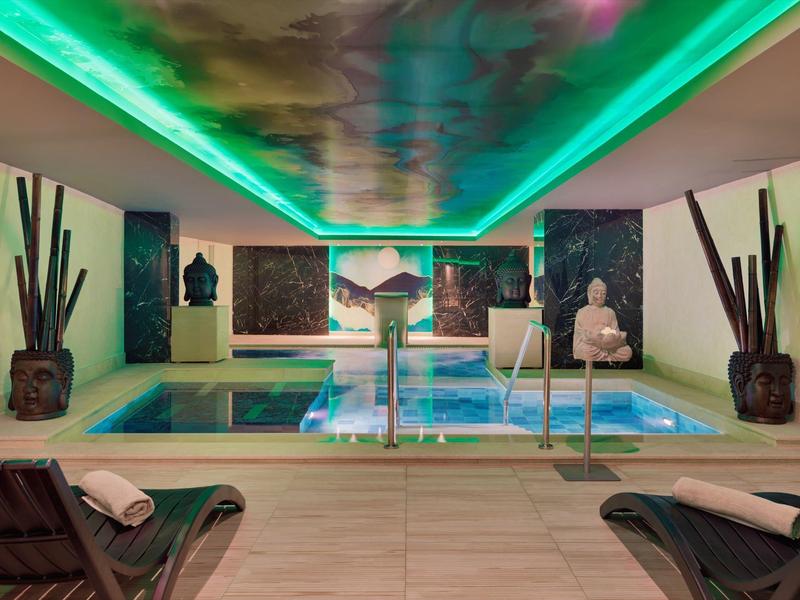 Moderner Wellnessraum mit beleuchtetem Hallenbad, grüner Deckenbeleuchtung und weißen Skulpturen.