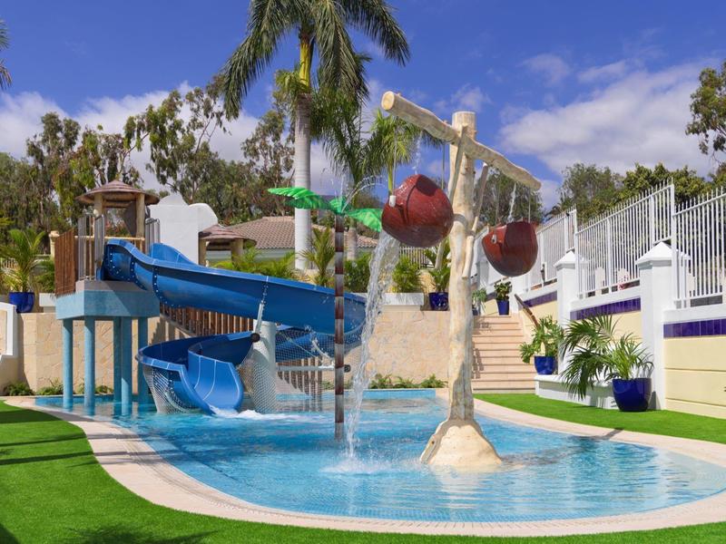 Wasserpark mit Rutschen, Wasserspielen, Palmen und bepflanztem Bereich bei klarem Himmel.