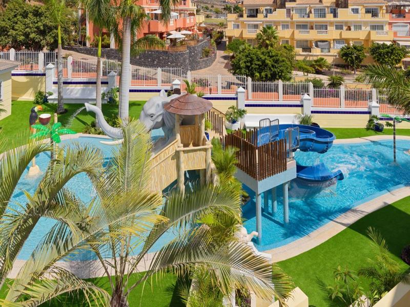 Wasserspielplatz mit Rutschen, mehreren Becken und grünem Rasen, umgeben von Wohngebäuden.