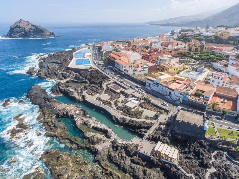 Littoral avec piscines rocheuses, une passerelle en mer et une ville aux toits rouges sous un ciel clair.