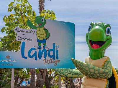 Grüne Schildkröte neben blauem Schild mit Text "Landi Village" und "Willkommen" im Freien.