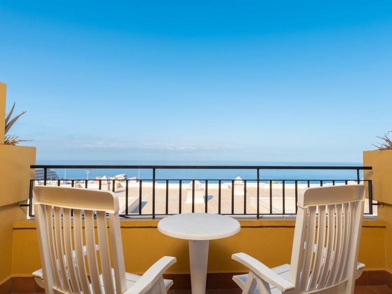 Balkon mit zwei weißen Stühlen und kleinem Tisch mit Meerblick unter blauem Himmel.