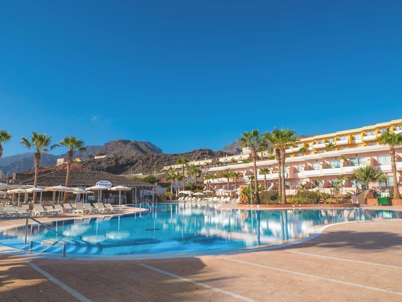 Großer Außenpool vor einem mehrstöckigen Hotel mit Palmen und Bergen im Hintergrund.