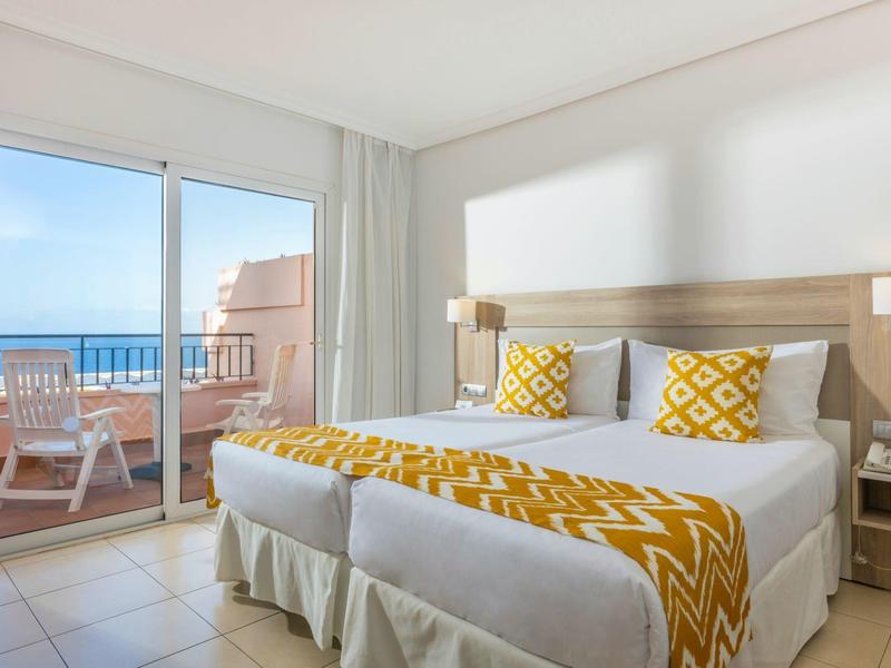 Helles Hotelzimmer mit zwei Betten, gelben Akzenten und Balkon mit Meerblick.
