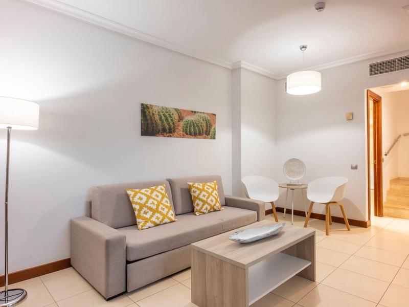 Helles, modernes Wohnzimmer mit grauem Sofa, gemusterten Kissen, Couchtisch und Essbereich in einem Ferienapartment.