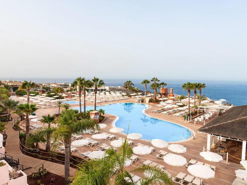 Hotelpool mit Sonnenliegen, Palmen und Blick auf das Meer am sonnigen Tag.