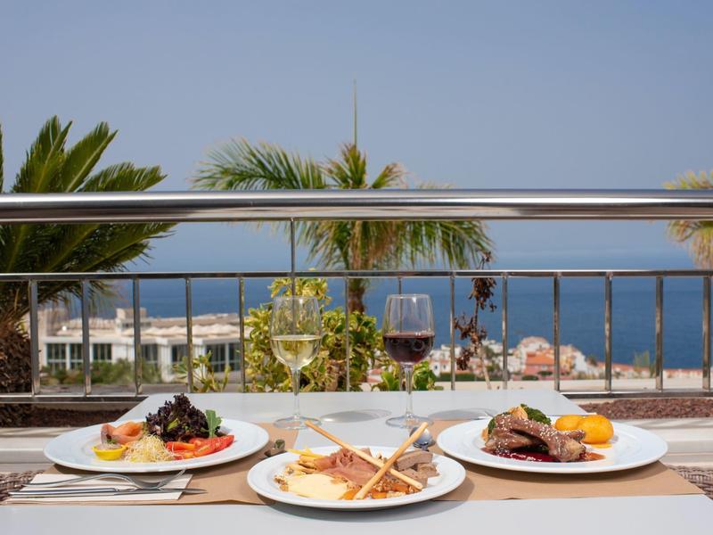 Drei Teller mit verschiedenen Speisen und ein Glas Rotwein auf einem Tisch mit Meerblick und Palmen.