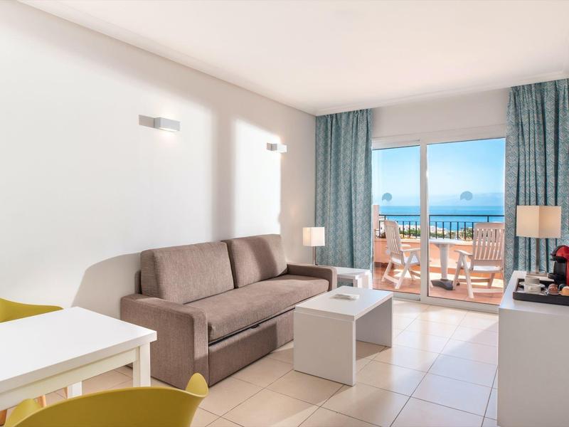 Helles Hotelzimmer mit Sofa, Tisch, Stühlen und Balkon mit Meerblick.