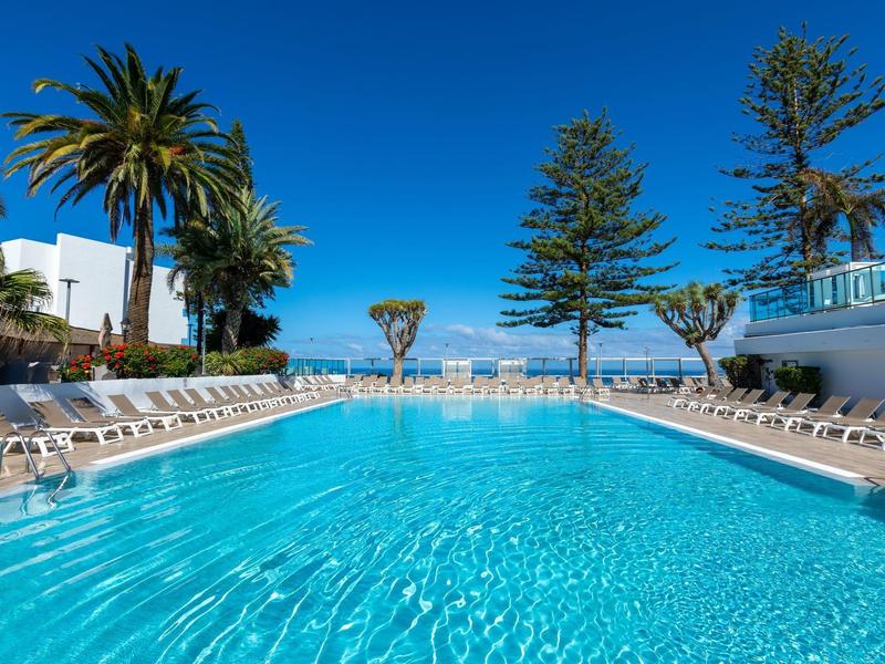 Gran piscina exterior con palmeras y tumbonas bajo un cielo azul despejado.