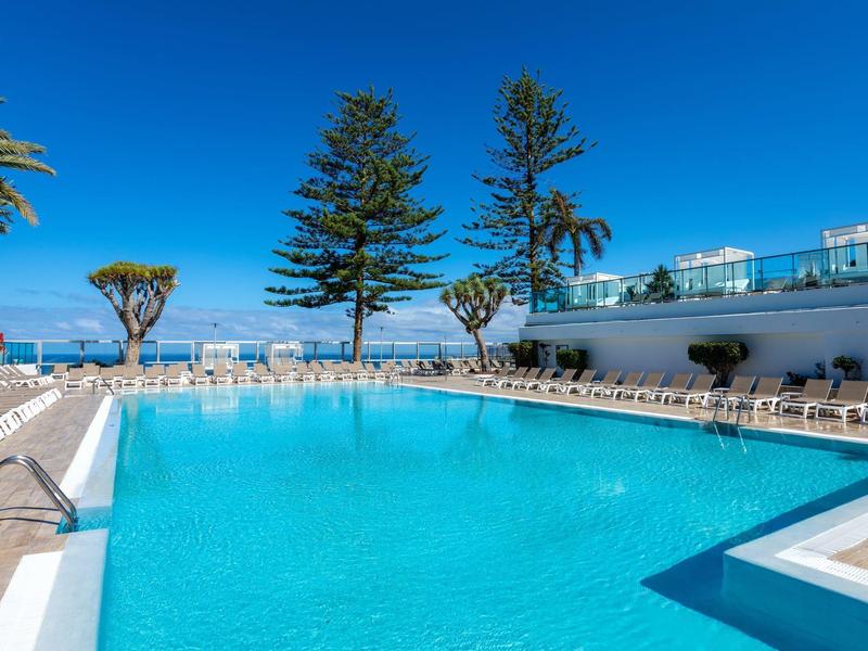 Piscina clara con tumbonas blancas y árboles altos bajo un cielo azul.