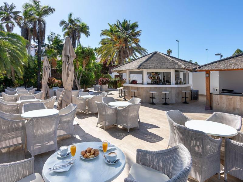 Terraza soleada de hotel con mesas blancas, sillas y un bar frente a palmeras.