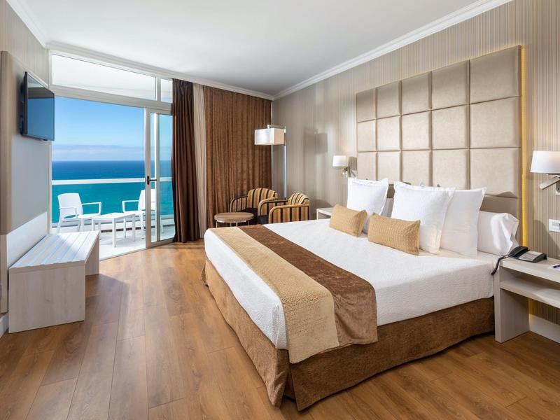 Habitación de hotel moderna con cama doble, balcón y vista al mar, suelos de madera claros y muebles cómodos.