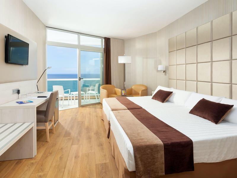 Habitación de hotel moderna con vista al mar, cama doble, escritorio y balcón con zona de estar.