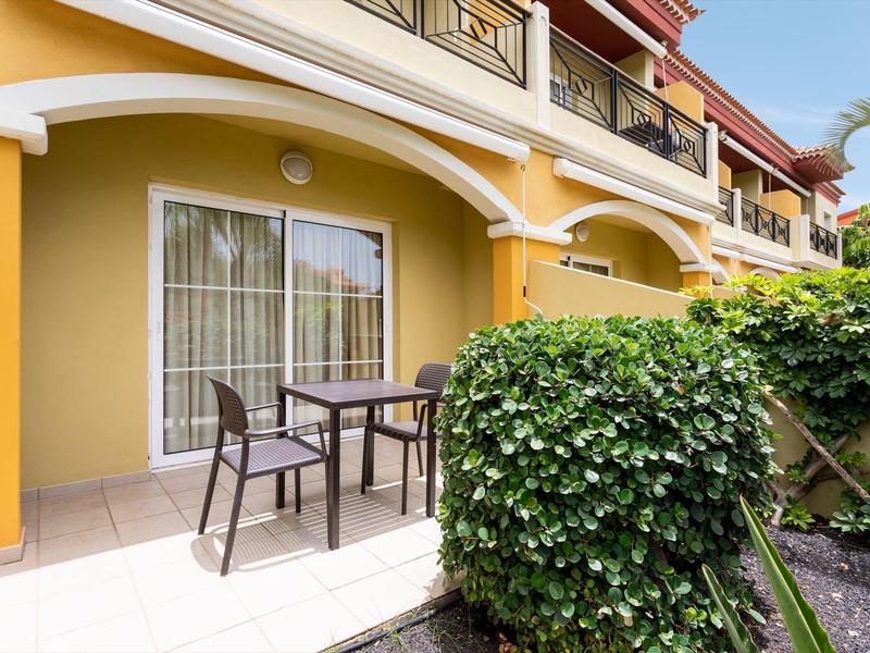 Terrasse avec table et deux chaises devant un bâtiment jaune avec balcons et haie verte.