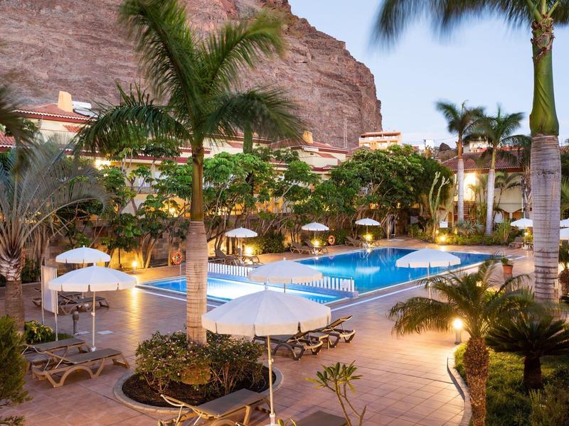 Zone de piscine éclairée avec palmiers et chaises longues dans une station tropicale au crépuscule.