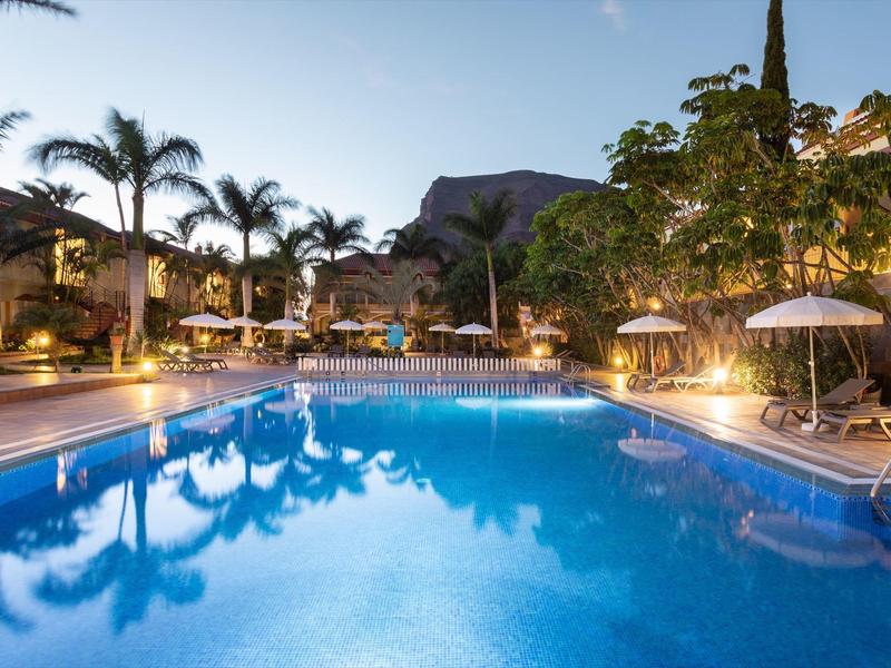 Piscine éclairée avec palmiers et chaises longues au crépuscule dans un complexe hôtelier tropical.
