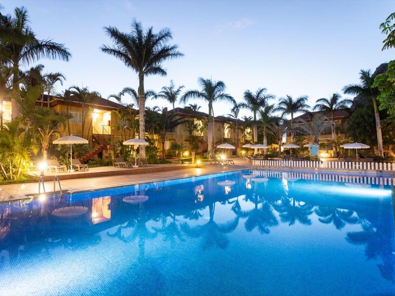 Piscine éclairée avec palmiers et chaises longues au coucher du soleil dans une station balnéaire.