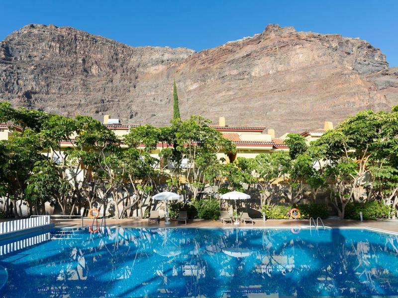 Piscine de loisirs avec eau claire bleue et jardin devant une montagne rocheuse sous un ciel bleu.