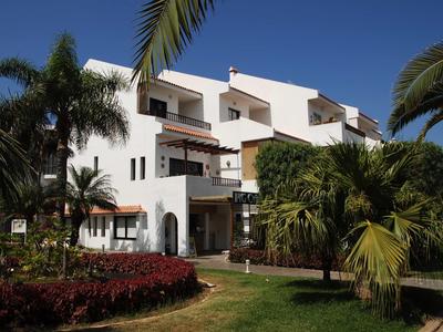 Bílá hotelová budova s několika balkony, obklopená palmami a zahradami pod jasnou oblohou.