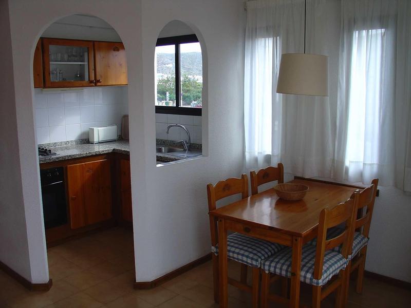 Kitchen and dining area with wooden chairs, table, and white curtains on the windows.