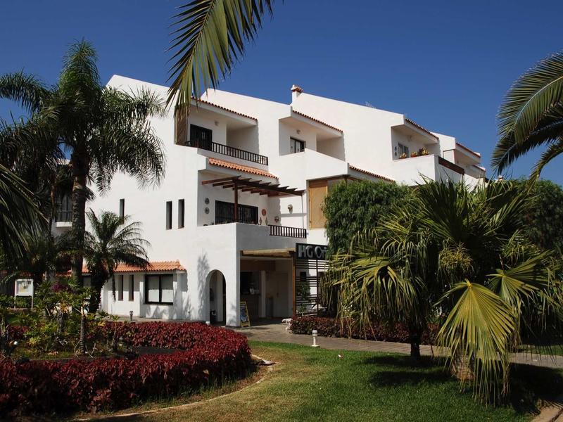 White hotel building with multiple balconies, surrounded by palm trees and well-maintained garden.