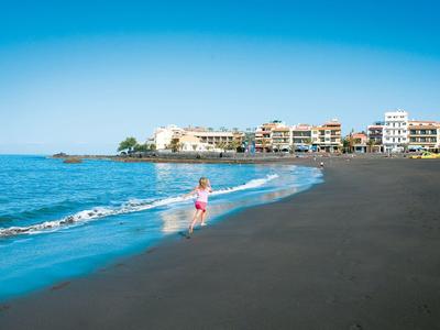 Plage de sable foncé, mer et un enfant jouant près des hôtels sous un ciel bleu.
