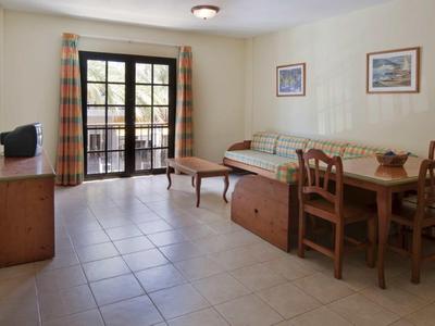 Lichte woonkamer met tegelvloer, bank, houten stoelen en grote ramen met gordijnen.