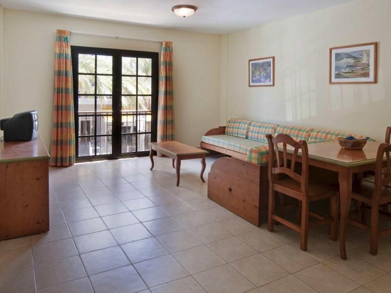 Lichte woonkamer met tegelvloer, bank, houten stoelen en grote ramen met gordijnen.