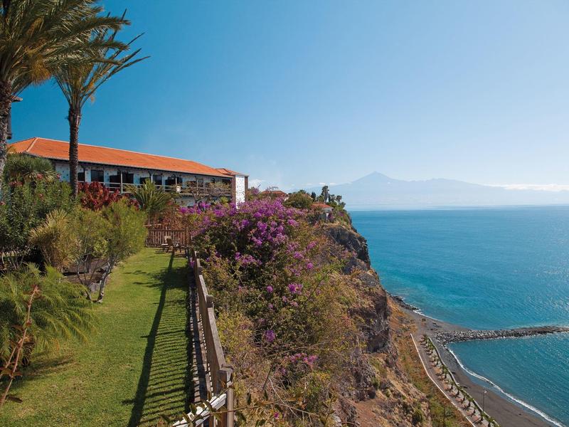 Hotel sulla scogliera con giardino e vista sul mare blu sotto un cielo limpido.