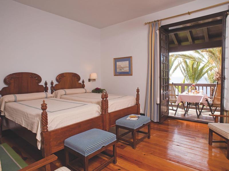 Camera d'hotel con letti singoli in legno, pavimento in legno e balcone con vista sulle palme.