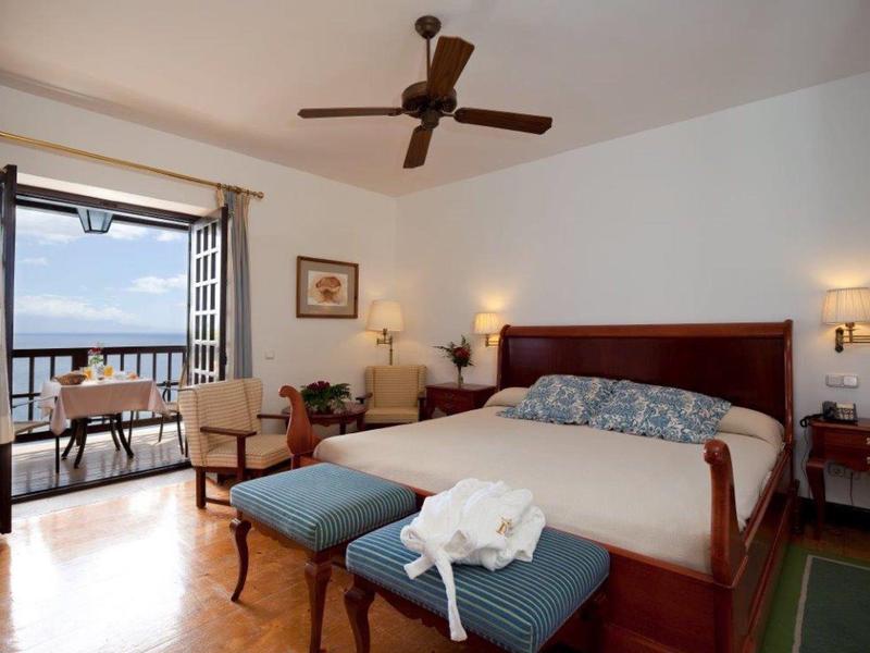 Camera d'hotel elegante con letto matrimoniale, poltrone, ventilatore a soffitto e balcone con vista mare.