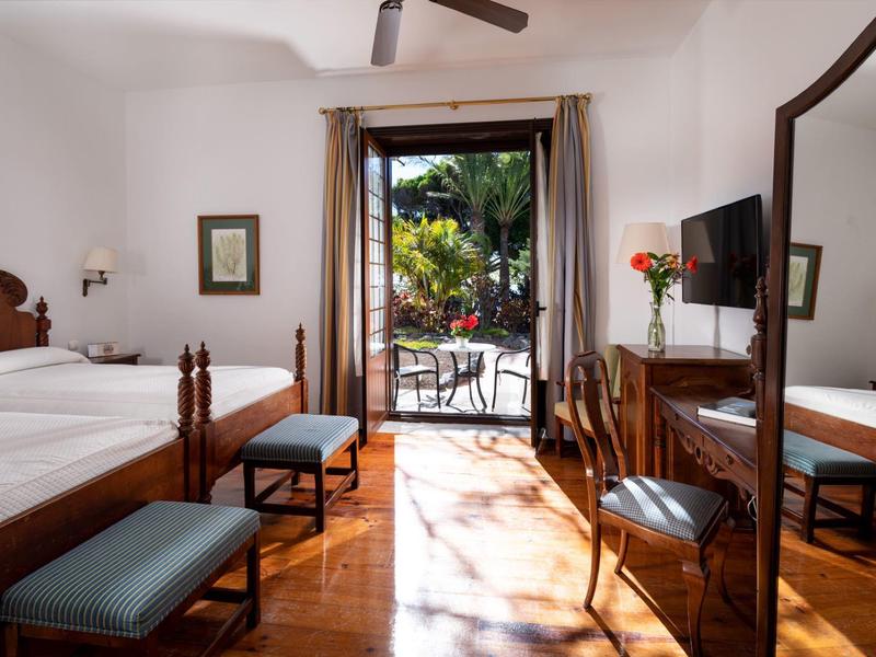 Camera d'hotel luminosa con letto matrimoniale, TV e vista su un giardino tropicale.